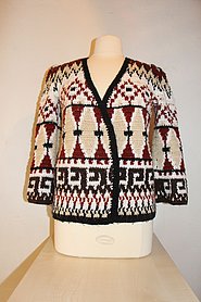 Fotoaufnahme einer Häkeljacke, die dem Torso einer Kleiderpuppe angezogen wurde. Die Häkeljacke ist im Folklorestil bzw. Ethno-Look gehalten und stammt aus den 1970ern. Der Kleiderpuppentorso steht auf einem Tisch aus Holz. 