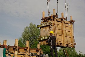 Manöver mit dem ersten Ganzteil Fotoaufnahme des Manövers mit dem ersten Ganzteil. Das Bauteil mit den Bretterwänden auf allen Seiten wird von einem Kran auf seine Position in der Außenwand gehoben. Ein Arbeiter mit Helm steht daneben und bringt es auf die richtige Stelle. Dahinter sind Baumkronen erkennbar.