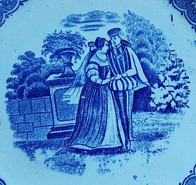 Auf dem Teller sind zwei Personen. Eine Frau und ein Mann. Sie halten sich an den Händen. Sie stehen in einem Park. Hinter ihnen ist ein Podest mit Blumenvase.