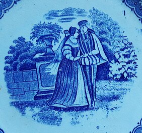 Auf dem Teller sind zwei Personen. Eine Frau und ein Mann. Sie halten sich an den Händen. Sie stehen in einem Park. Hinter ihnen ist ein Podest mit Blumenvase.