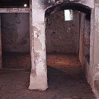 FFM_Mittelalter_Matting_Durchgaenge Fotoaufnahme der Innenräume im Erdgeschoss des Steinhauses aus Matting am aktuellen Standort. Im Raum befinden sich mehrere Türen und Durchgänge. An der Wand rechts im Bild hängt eine Informationstafel, der Boden besteht aus gestampftem Lehm.