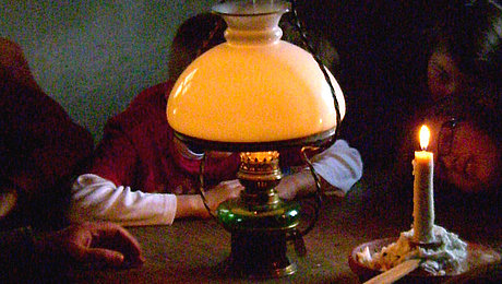 FFM_Feuer_und_Licht_im_Haus Fotoaufnahme der Führung „Feuer und Licht im Haus“. Um einen Holztisch sitzt eine Gruppe Kinder. In der Mitte stehen eine leuchtende Petroleumlampe und eine angezündete Kerze. Abgesehen von diesen schwachen Lichtquellen ist der Raum dunkel.