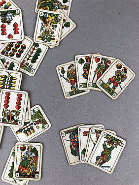 Auf dem Bild liegen viele Spielkarten. Sie zeigen Eicheln, Schellen, Herzen und Blätter. Die Figuren sehen aus wie Bauern, Soldaten und Könige.