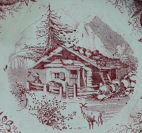 Auf dem Teller ist eine Holzhütte. Sie steht im Gebirge. Vor der Hütte steht eine Ziege. Hinter der Hütte steht eine Fichte.