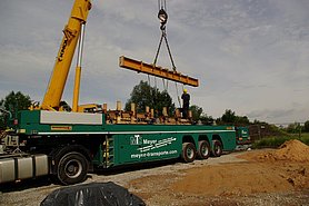 Fotoaufnahme eines Schwerlastkrans zum Heben der schweren Wandscheiben. Der Kran steht auf der Baustelle, auf seiner Ladefläche liegt ein mit Holzbrettern ummanteltes Mauerstück. Daneben steht ein Arbeiter mit Schutzhelm, der die Ketten des Krans am Bauteil befestigt. 