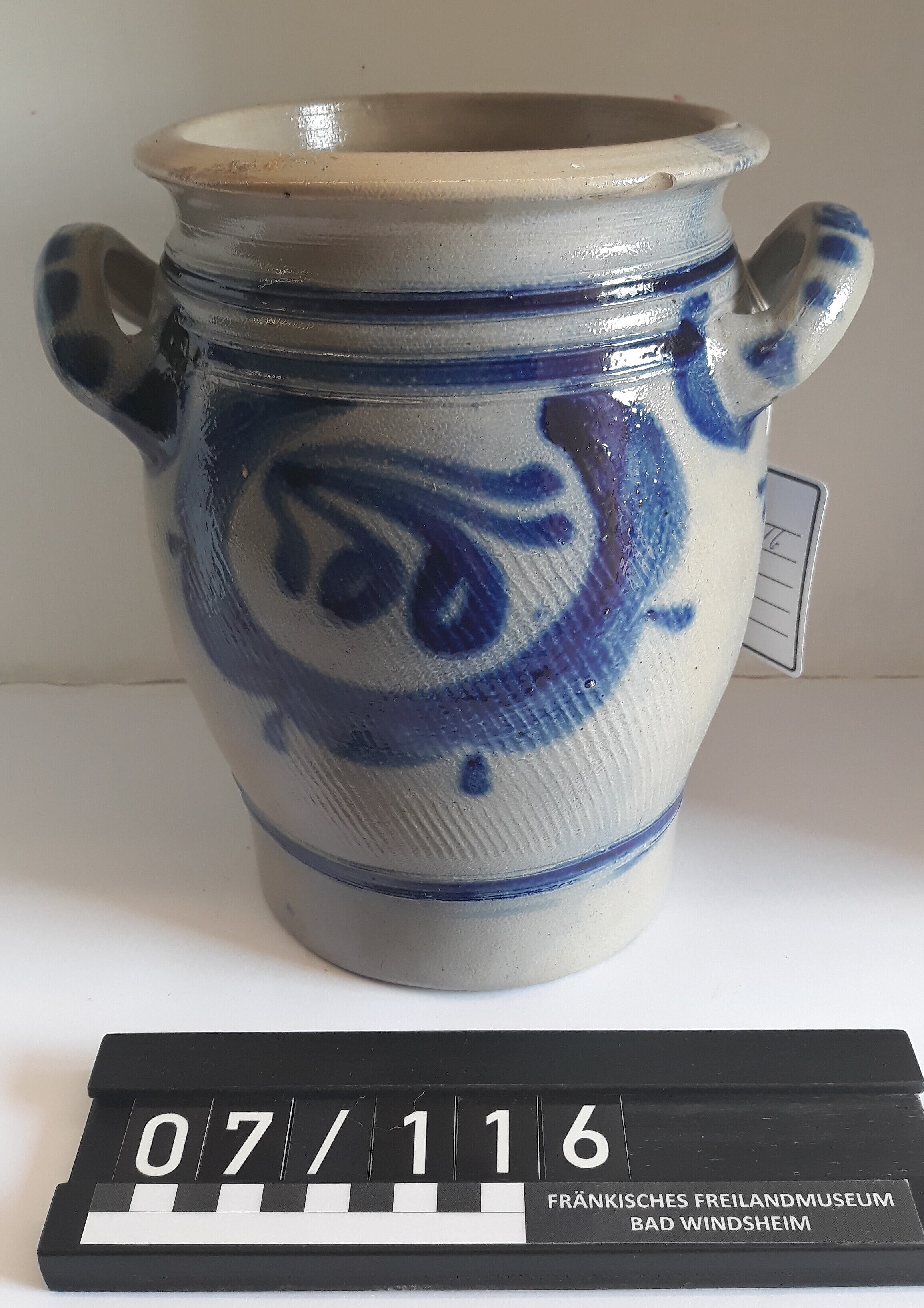 Der Topf ist grau. Er hat zwei Henkel. Auf dem Topf sind blaue Muster.