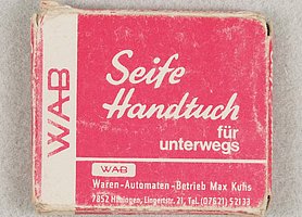 Die Schachtel ist rechteckig. Sie ist aus Pappe. Sie ist rot bedruckt. Die Schrift ist weiß. in der Schachtel steckt ein Tuch.