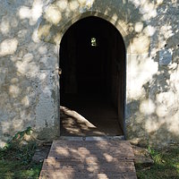 FFM_Eingangsschwelle_Matting Fotoaufnahme des Eingangs vom Steinhaus aus Matting am aktuellen Standort. Auf den Eingangsbereich fällt der Schatten eines Baumes. Zur nach innen geöffneten Holztür führt eine Steinstufe, vor der Stufe und der Türschwelle liegt je eine Rampe aus Holz. Links und rechts davon wächst Gras.