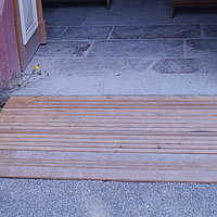 FFM_West_Kapelle_Rampe Fotoaufnahme des Eingangs der Kapelle aus Rodheim am aktuellen Standort. Vor der Schwelle liegt eine Rampe aus Holz. Der Boden im Inneren besteht aus Steinplatten. Neben der Tür blühen Wildblumen.