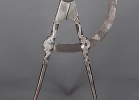 Der Zirkel ist aus Eisen. Er hat zwei Schenkel. Beide sind vorne spitz. Sie sind miteinander verbunden. Sie können an einem Bogen entlang gedreht werden. Die Schenkel sind verziert.