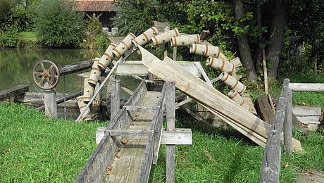 Fotoaufnahme des hölzernen Wasserschöpfrades. Das stillstehende Rad befindet sich teilweise im Wasser und ist mit Wasserkübeln besetzt. Wenn das Rad in Betrieb ist, kippen die Kübel das Wasser in eine Holzrinne, die vor dem Rad auf einer Wiese aufgebaut ist. Am Ufer stehen einige Bäume. 
