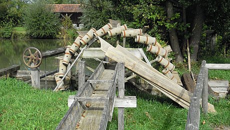 FFM_Wasserschoepfrad_Internet Fotoaufnahme des hölzernen Wasserschöpfrades. Das stillstehende Rad befindet sich teilweise im Wasser und ist mit Wasserkübeln besetzt. Wenn das Rad in Betrieb ist, kippen die Kübel das Wasser in eine Holzrinne, die vor dem Rad auf einer Wiese aufgebaut ist. Am Ufer stehen einige Bäume.