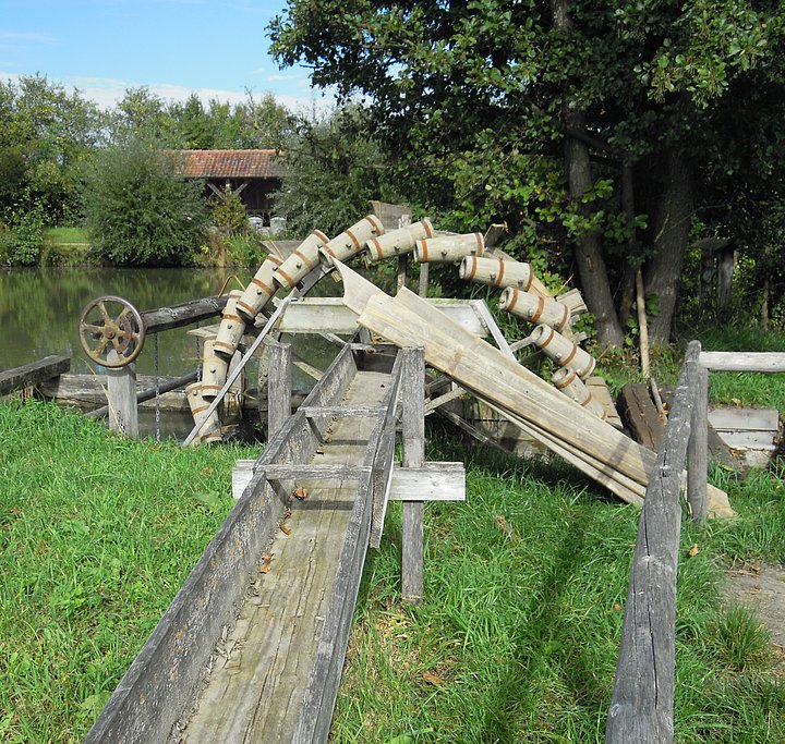 FFM_Wasserschoepfrad_Internet Fotoaufnahme des hölzernen Wasserschöpfrades. Das stillstehende Rad befindet sich teilweise im Wasser und ist mit Wasserkübeln besetzt. Wenn das Rad in Betrieb ist, kippen die Kübel das Wasser in eine Holzrinne, die vor dem Rad auf einer Wiese aufgebaut ist. Am Ufer stehen einige Bäume.