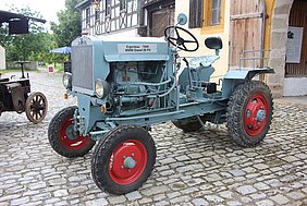 Der Schlepper ist hellblau. Die Räder sind rot. Er steht im Museum.