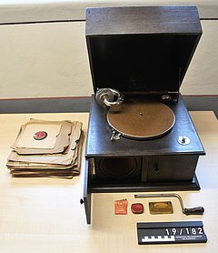Das Grammophon ist kistenförmig. Der Deckel ist geöffnet. Daneben liegen eine Kurbel und Ersatznadeln. Mehrere Schellackplatten können abgespielt werden. Sie enthalten Schlagermusik.