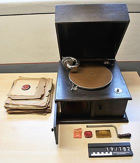 Das Grammophon ist kistenförmig. Der Deckel ist geöffnet. Daneben liegen eine Kurbel und Ersatznadeln. Mehrere Schellackplatten können abgespielt werden. Sie enthalten Schlagermusik.