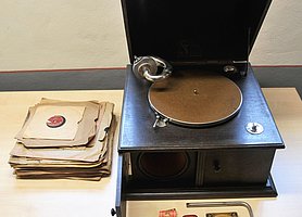 Das Grammophon ist kistenförmig. Der Deckel ist geöffnet. Daneben liegen eine Kurbel und Ersatznadeln. Mehrere Schellackplatten können abgespielt werden. Sie enthalten Schlagermusik.