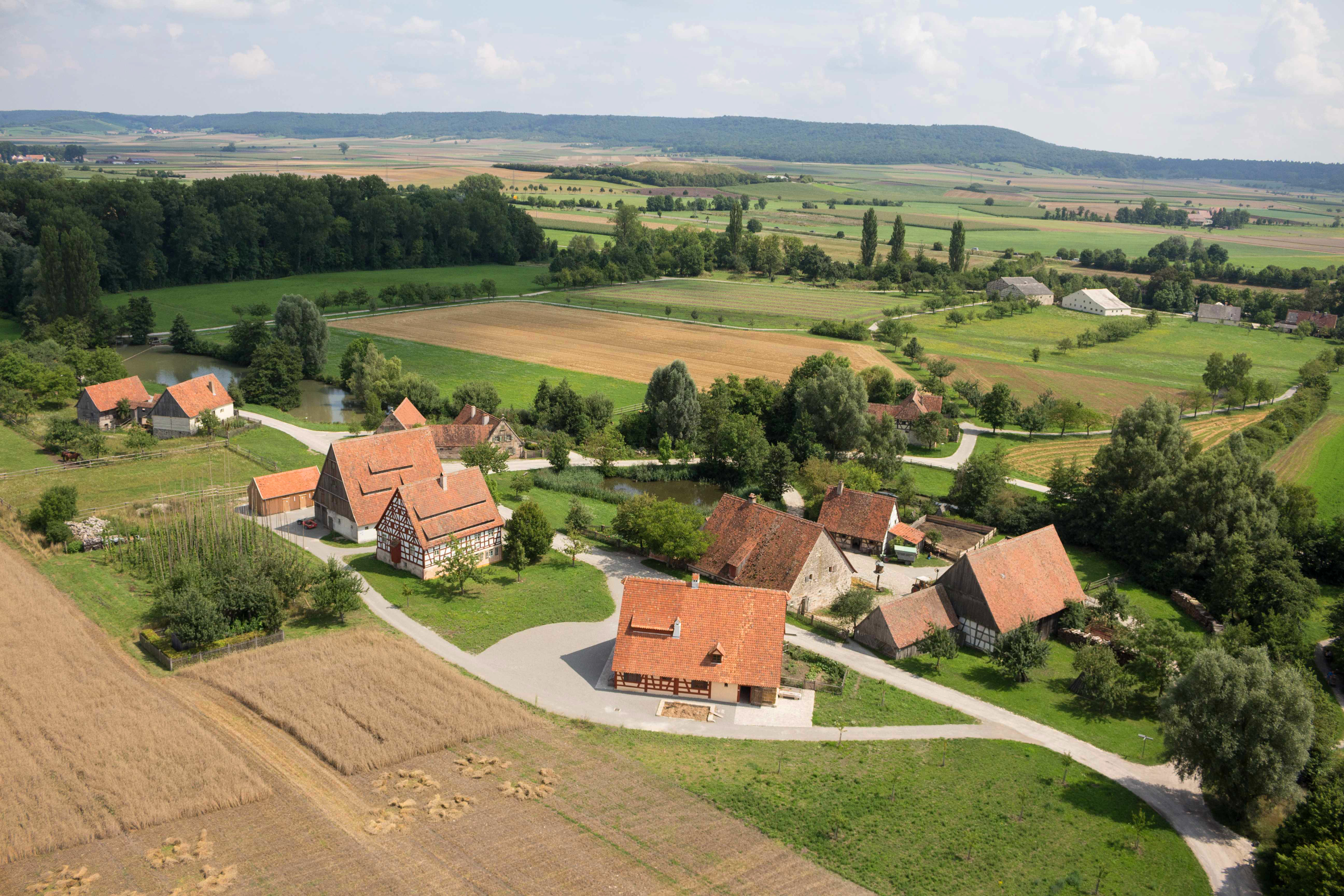 FFM_Baugruppe_Ost Fotoaufnahme eines Luftbildes der Baugruppe Ost. Die Häuser stehen in kleinen Gruppen und sind umgeben von Feldern, Wiesen und Bäumen. Im Hintergrund ist die weite Landschaft des Museums und der Umgebung erkennbar.