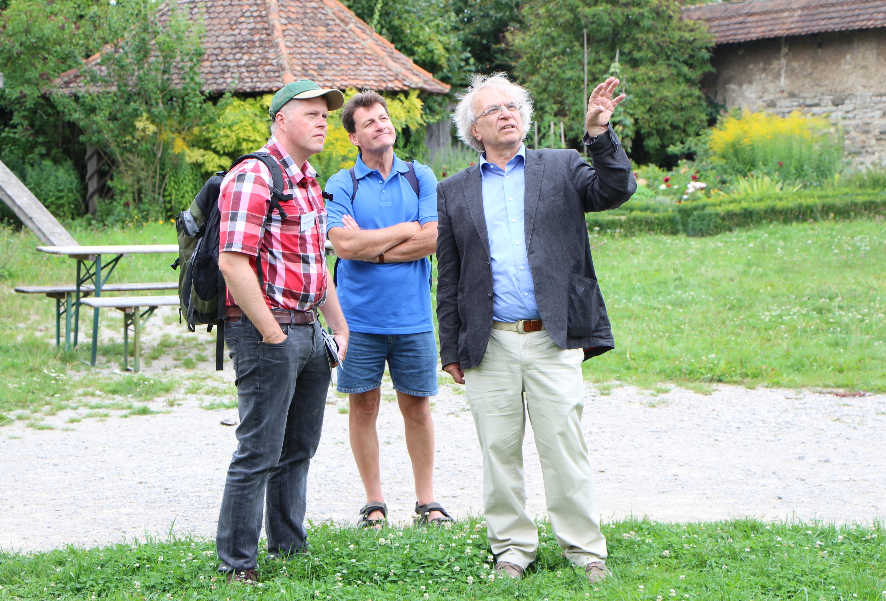 FFM_Fuehrungen Fotoaufnahme zweier Besucher mit einem Museumsführer. Die drei Männer betrachten etwas außerhalb des Bildes. Einer steht auf dem Kiesweg, die anderen beiden im Gras. Im Hintergrund befinden sich eine Bierzeltgarnitur sowie Büsche, Blumen und Gebäude.