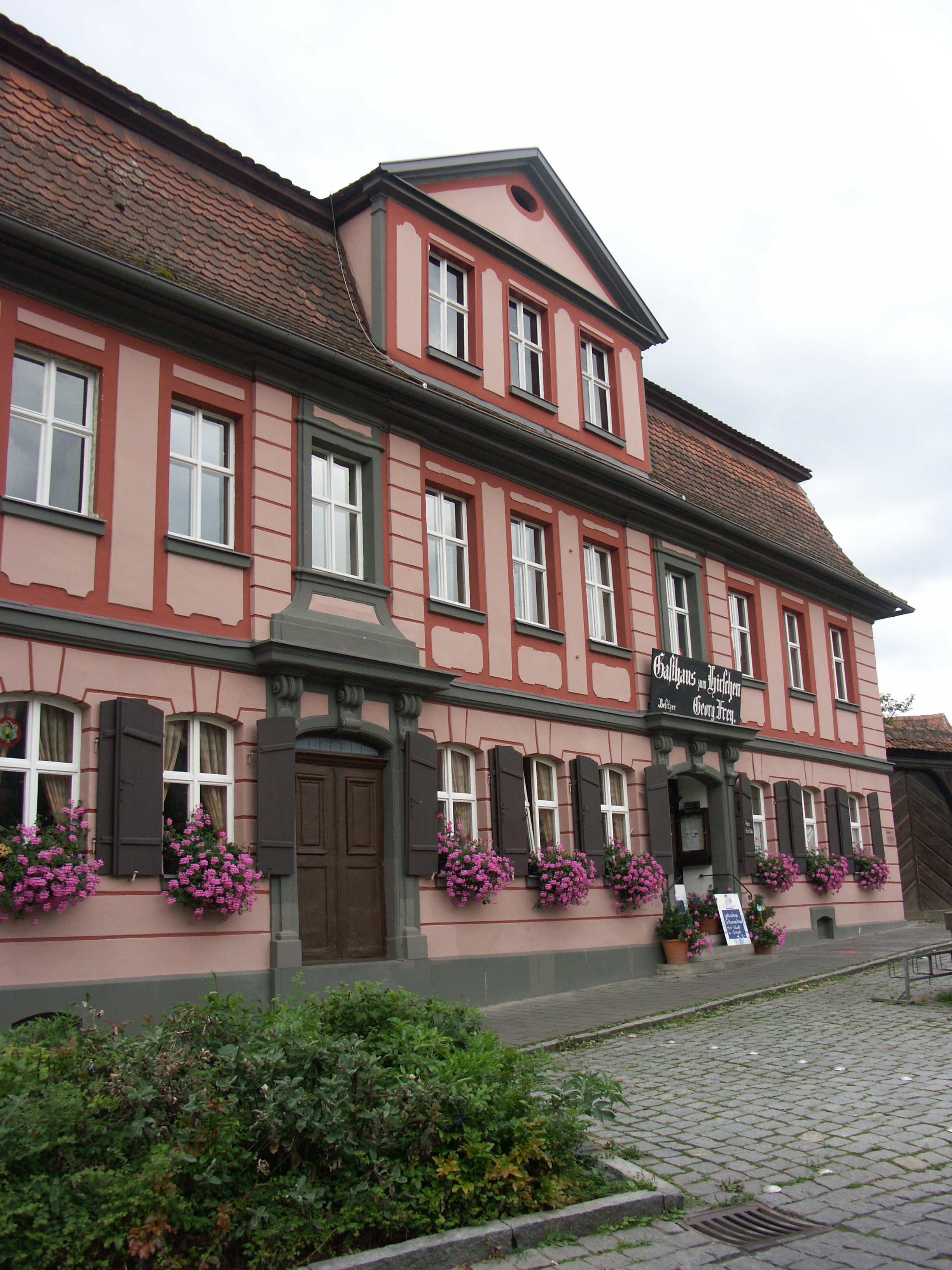 Ein repräsentatives, zweigeschossiges Gasthaus, rosa gestrichener Putz mit roten Fenstereinfassungen. Im Erdgeschoss zieren die Fensterkästen lila Blumen