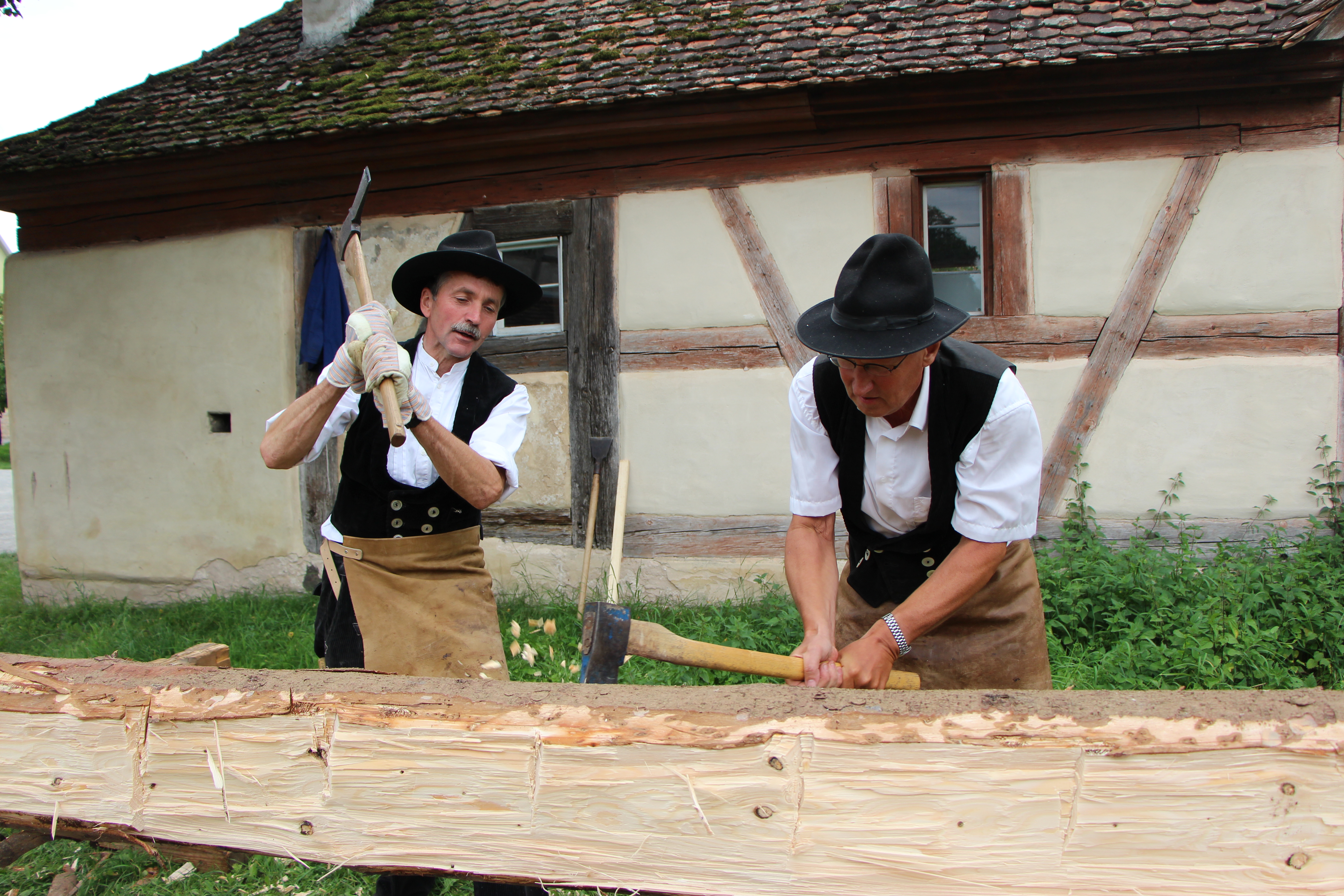 Fotoaufnahme vom Handwerkertag. Gezeigt werden zwei Zimmermännern in Kluft, die einen aufgebockten Holzstamm mit Beilen bearbeiten. Im Hintergrund steht ein mit Flachziegeln gedecktes Fachwerk. 
