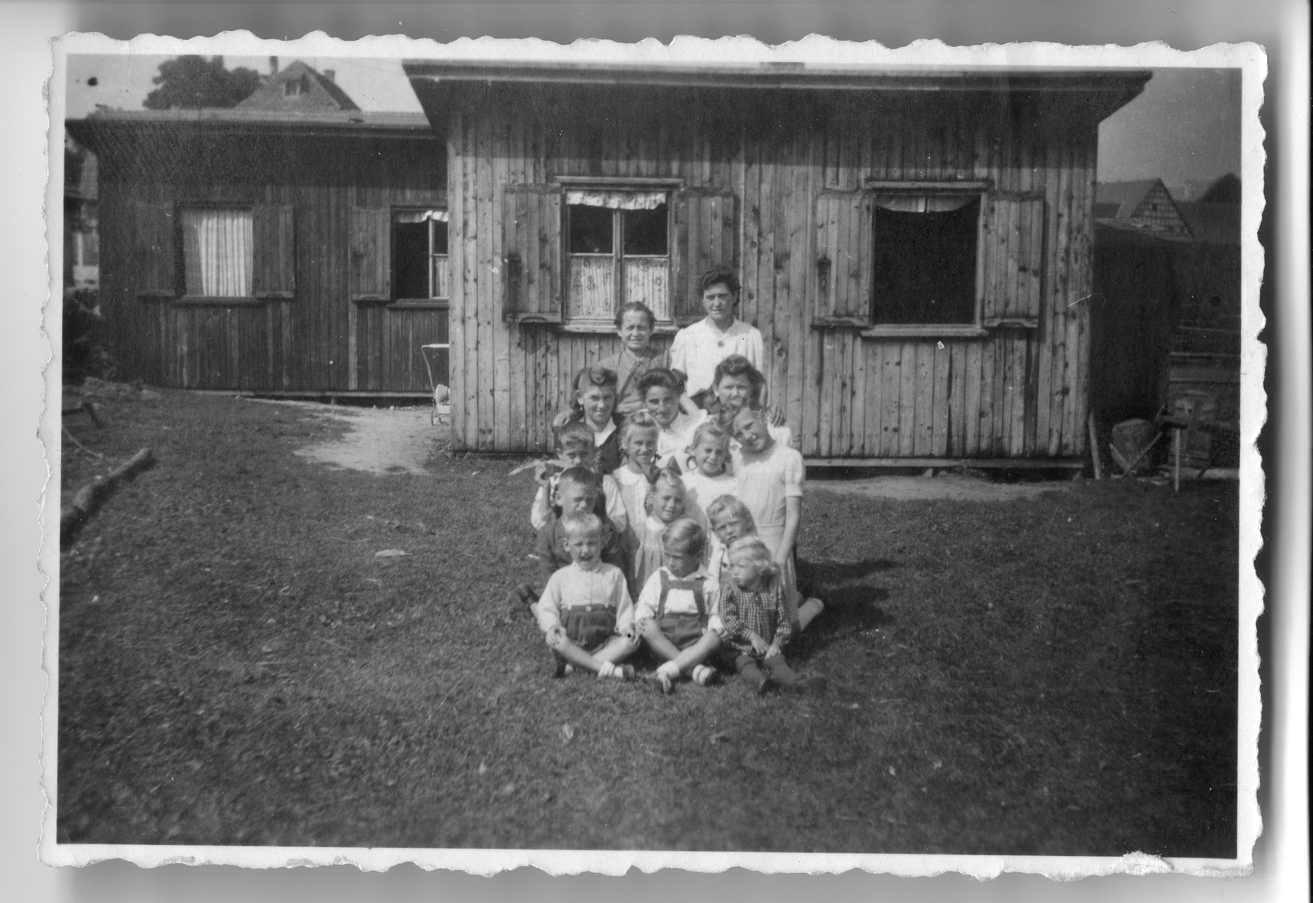 Die beiden Behelfsheime sind kleine Holzhäuser. Davor stehen Frauen und Kinder. Sie wohnen in den Häusern.