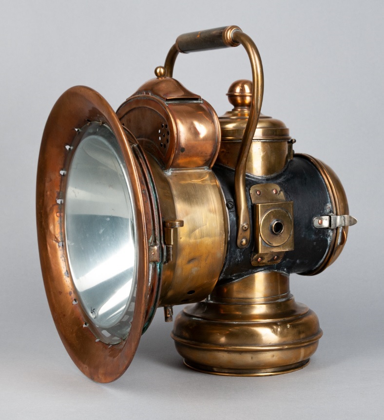 Die Lampe ist aus Messing. Sie hat einen Griff. Nach vorne sieht sie aus wie ein Trichter. Der Trichter verteilt das Licht.