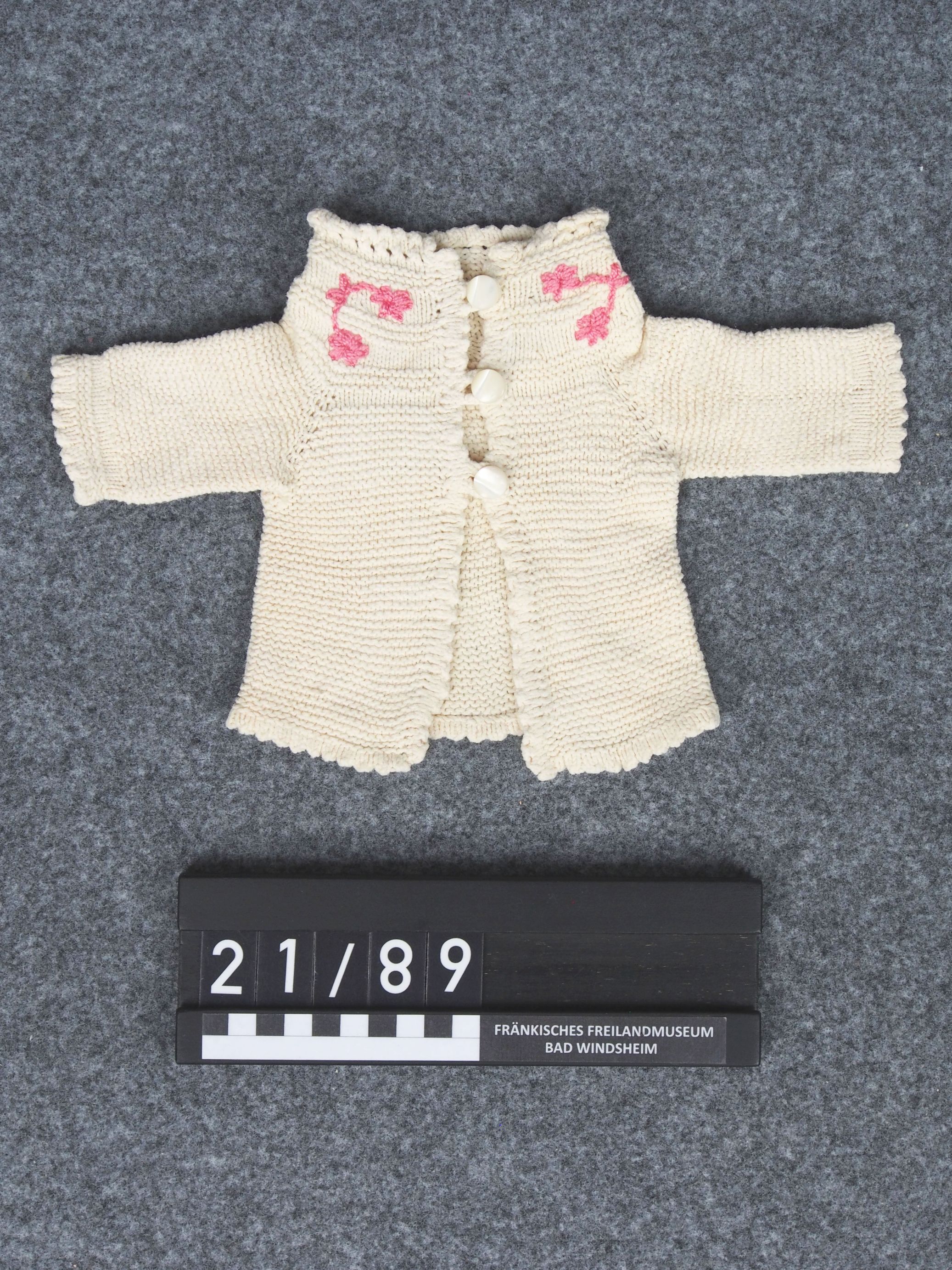 Die kleine Jacke ist gestrickt. Sie ist beige. Auf dem Kragen sind rosafarbene Blumen.