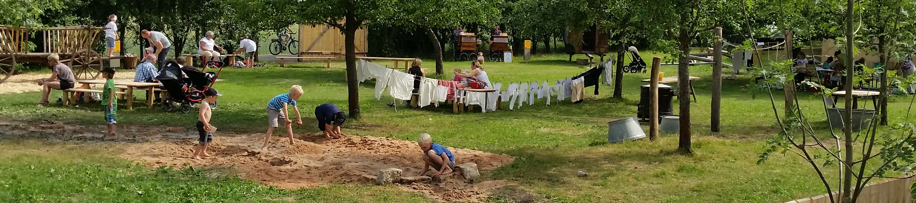 Fotoaufnahme des Erlebnisplatzes für Kinder in der Baugruppe Süd. In der Mitte des Bildes spielen einige Jungen im Sand. Dahinter hängen zwei Wäscheleinen, die zwischen Bäumen gespannt wurden. An einem Holztisch auf der linken Seite sitzen Eltern und schauen ihren Kindern beim Spielen zu. 