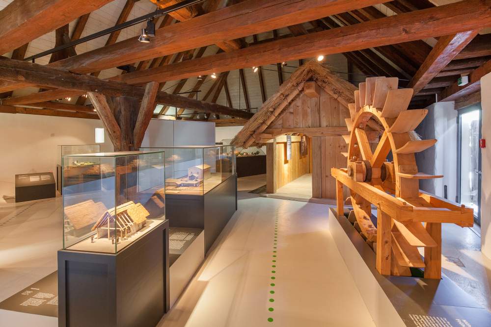 FFM_Ausstellung_Archaeologische_Staatssammlung Fotoaufnahme des Innenraums vom Archäologiemuseum in der Schafscheune aus Virnsberg. Ein Bodenleitsystem führt durch die Ausstellung. Links vom Weg stehen Vitrinen mit Modellen. Rechts ein hölzerner Wasserrad. Dahinter ist eine kleine Hütte aus Holz aufgebaut.